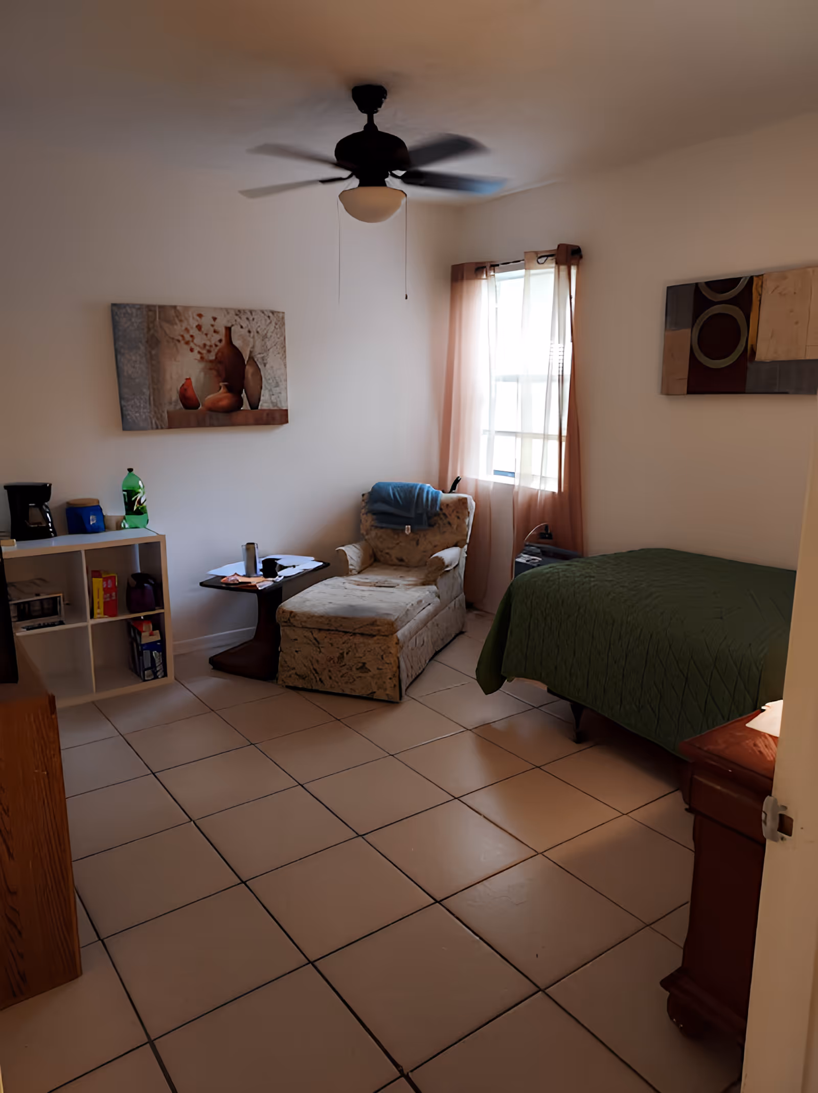A cozy bedroom with a single bed covered in a green quilt, a cushioned armchair with a blue blanket draped over it, a small side table with various items, a white shelving unit holding a coffee maker and other items, two pieces of wall art, a window with sheer curtains, and a ceiling fan overhead.