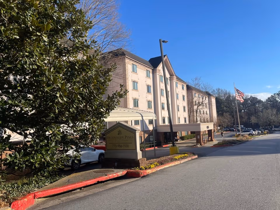 Front exterior of Dunwoody Pines Retirement Community showing the building entrance, driveway, sign, parked cars, and an American flag.