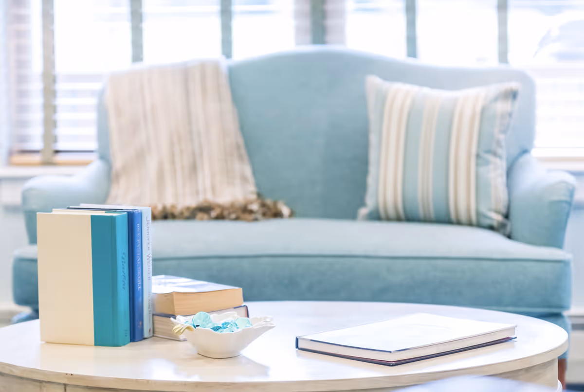 A light blue sofa with a striped pillow and a beige throw blanket in the background. In front of the sofa is a round white coffee table with a stack of books, a small decorative bowl with blue glass stones, and an open book.