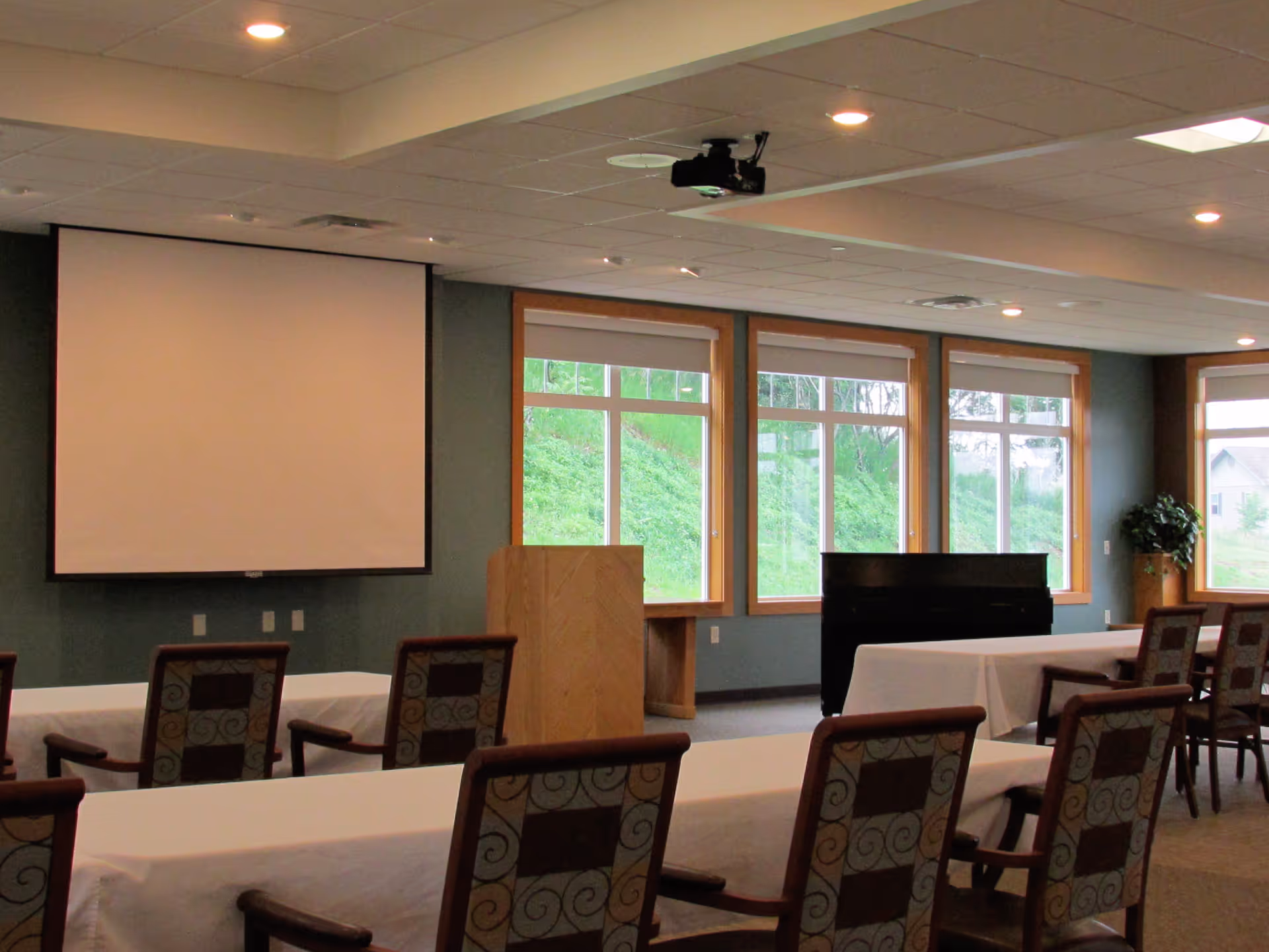 Conference/activity room with tables and chairs facing a projection screen and podium in front of large windows.