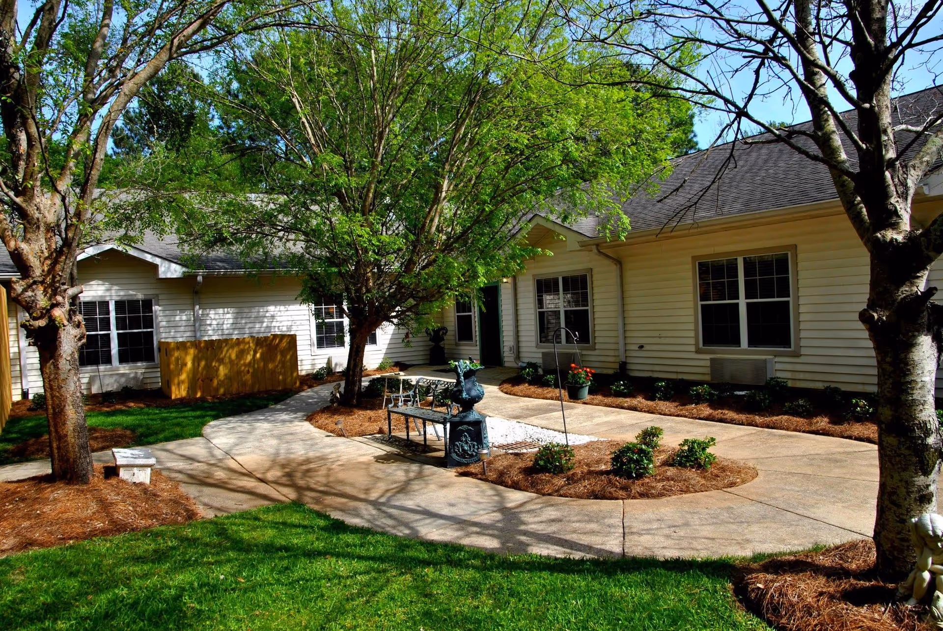Courtyard with a paved walkway, trees, benches and a small fountain in front of a single-story assisted living building.