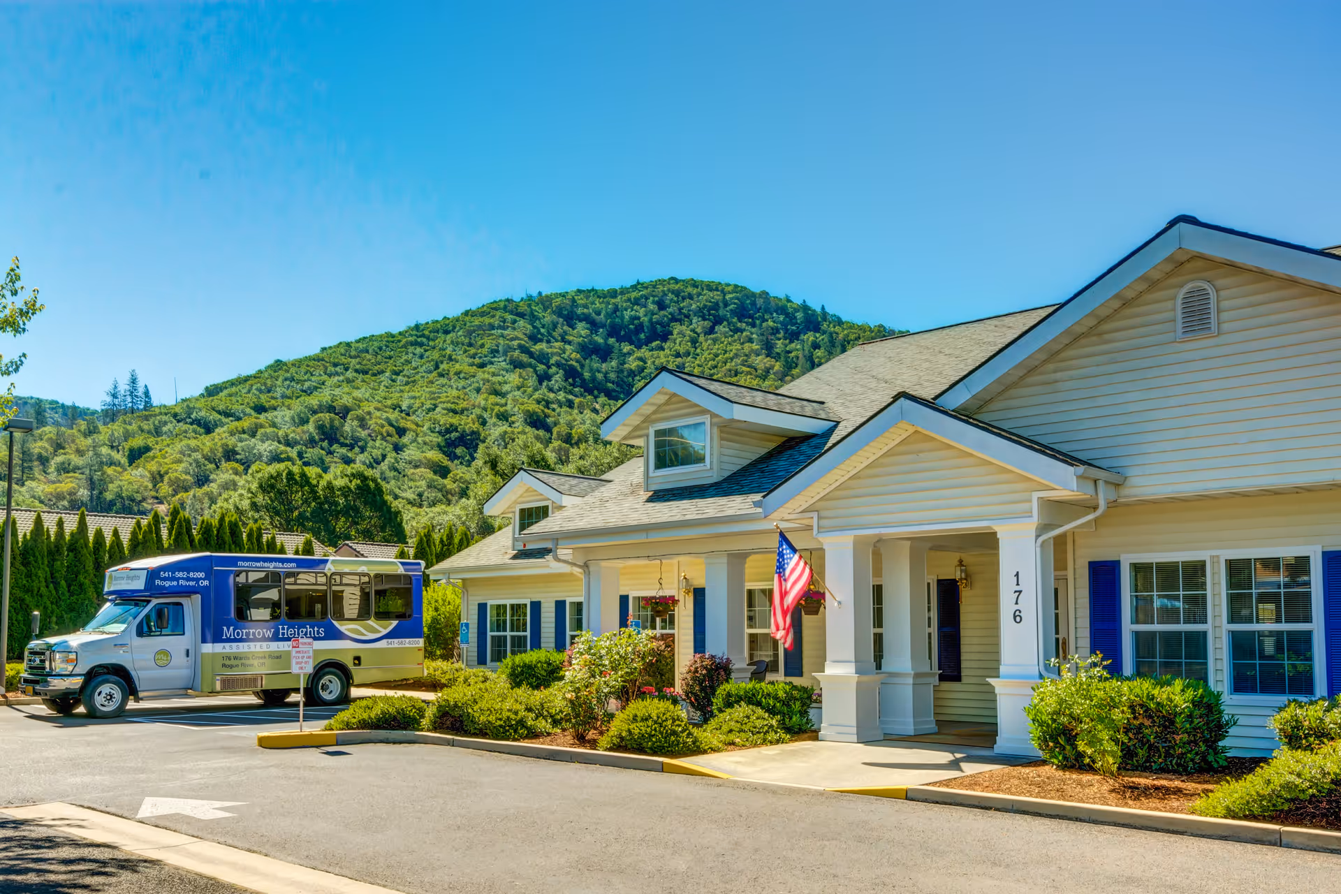 Exterior view of Morrow Heights Assisted Living facility with a single-story building featuring white siding, blue shutters, and a covered entrance with an American flag. A shuttle bus with Morrow Heights branding is parked nearby. The background shows a green, tree-covered hill under a clear blue sky.
