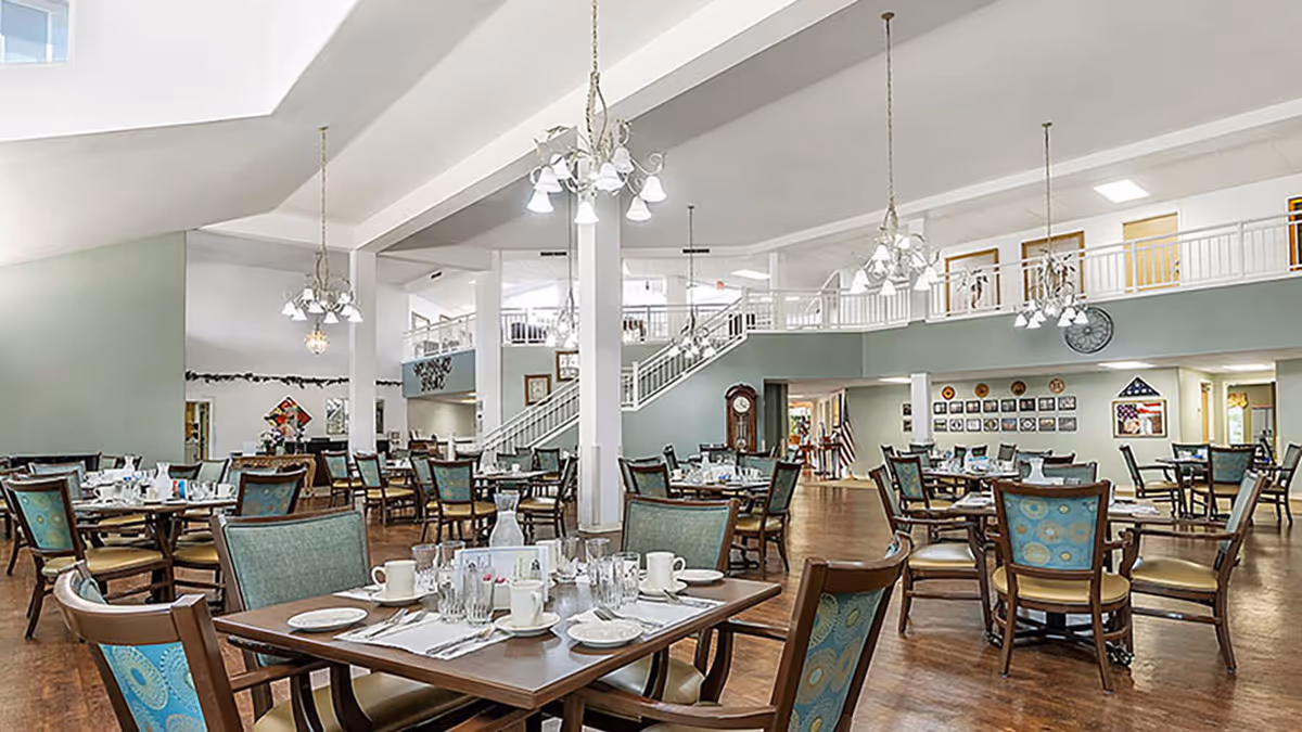 Spacious senior living dining room with multiple set tables and chairs, high ceilings, chandeliers, and an open second-floor balcony.
