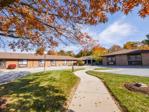 Exterior view of a senior living facility with brick wings, a curved walkway, and fall foliage.
