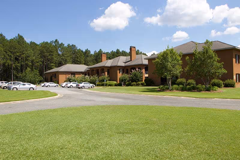 Brick senior living facility building with a circular driveway, parked cars, and a manicured lawn under a blue sky.