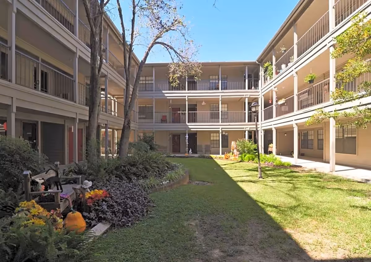 Outdoor courtyard area of a senior living facility with a grassy lawn, trees, plants, and flowers. The courtyard is surrounded by a three-story building with balconies and railings. There is a bench with decorative items and a lamppost on the lawn under a clear blue sky.