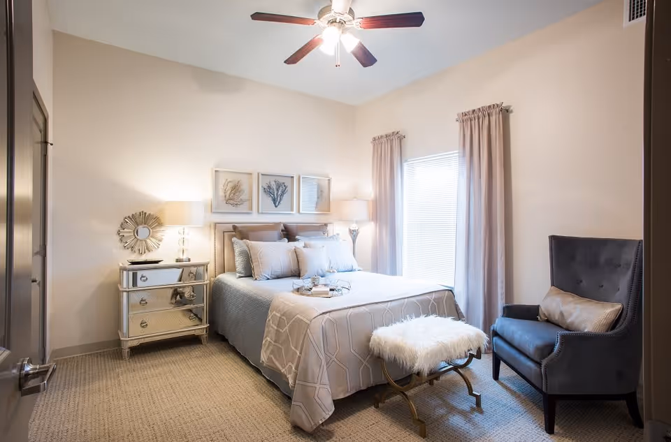 A well-lit bedroom with a neatly made bed featuring multiple pillows and a light-colored bedspread. There is a mirrored nightstand with a lamp and decorative mirror on the left side of the bed. Above the bed are three framed botanical prints. A window with light curtains is on the right wall, next to a dark upholstered armchair with a decorative pillow. A small bench with a furry white cushion is placed at the foot of the bed. The ceiling has a fan with lights.