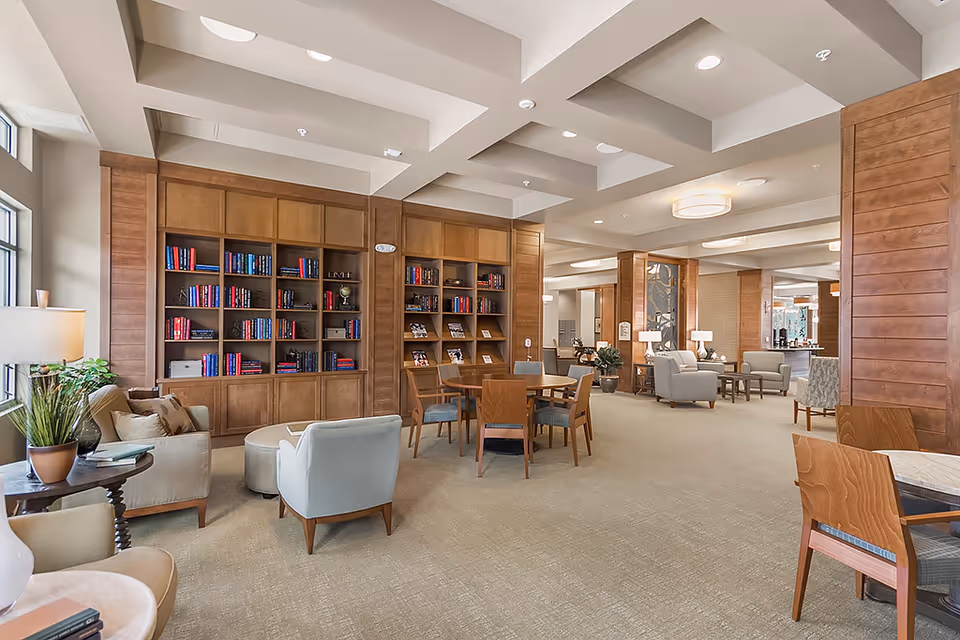 Spacious and well-lit senior living common area with beige carpet, wooden bookshelves filled with books, multiple seating arrangements including armchairs, sofas, and wooden chairs around tables, and decorative plants and lamps. The ceiling has recessed lighting and modern light fixtures.