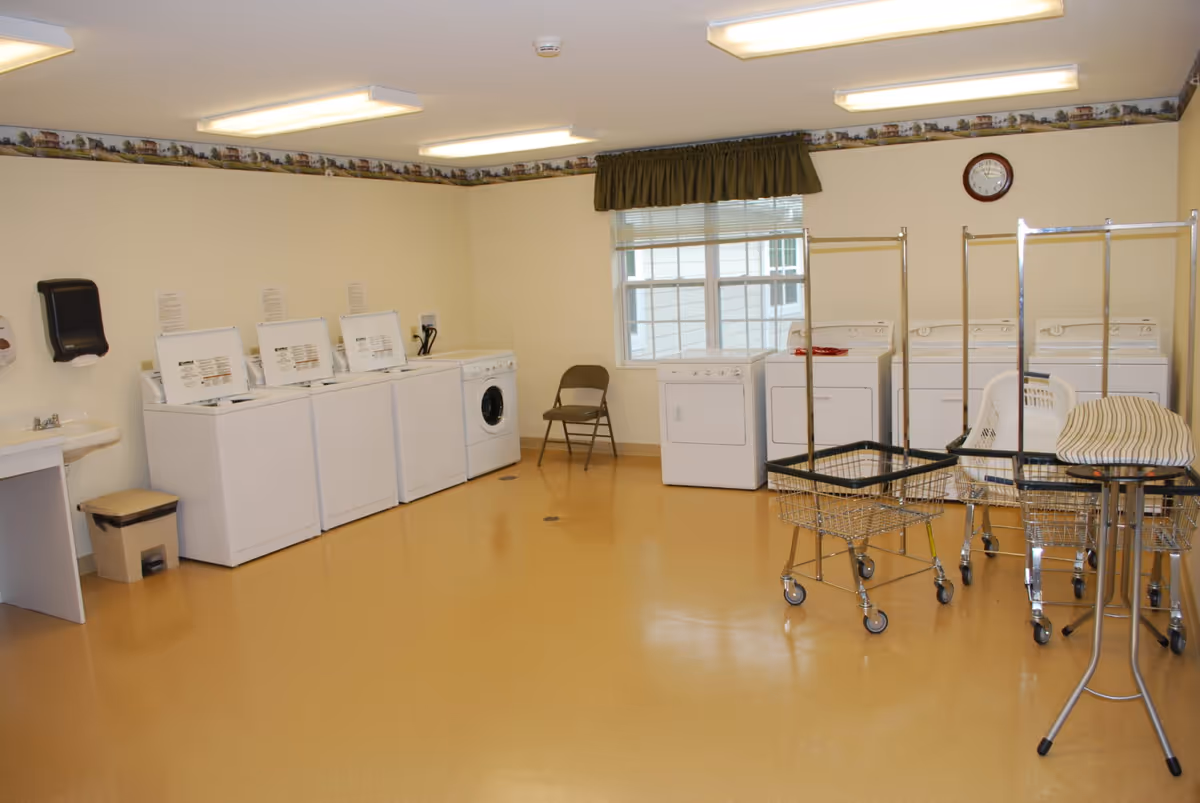 A laundry room with multiple washing machines and dryers along the walls. There is a folding chair near a window with blinds and a green valance. Several laundry carts and an ironing board are positioned in the room. The floor is a light brown color, and the walls are cream with a decorative border near the ceiling. A wall clock is mounted above the dryers.