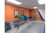 Interior rehabilitation gym with parallel bars, exercise balls, and wall-mounted equipment along orange and blue walls.