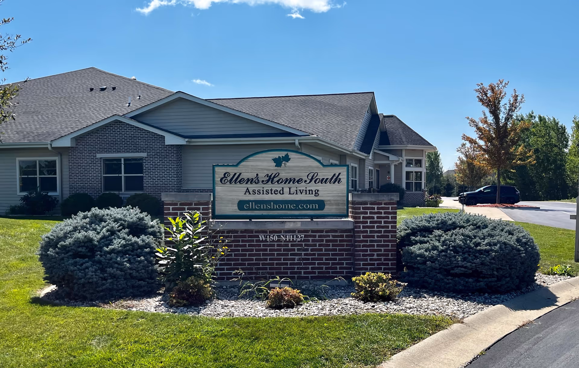 Exterior view of Ellen's Home South Assisted Living facility showing a brick sign with the facility name and website, surrounded by landscaped bushes and grass, with the building and a parking lot in the background under a clear blue sky.