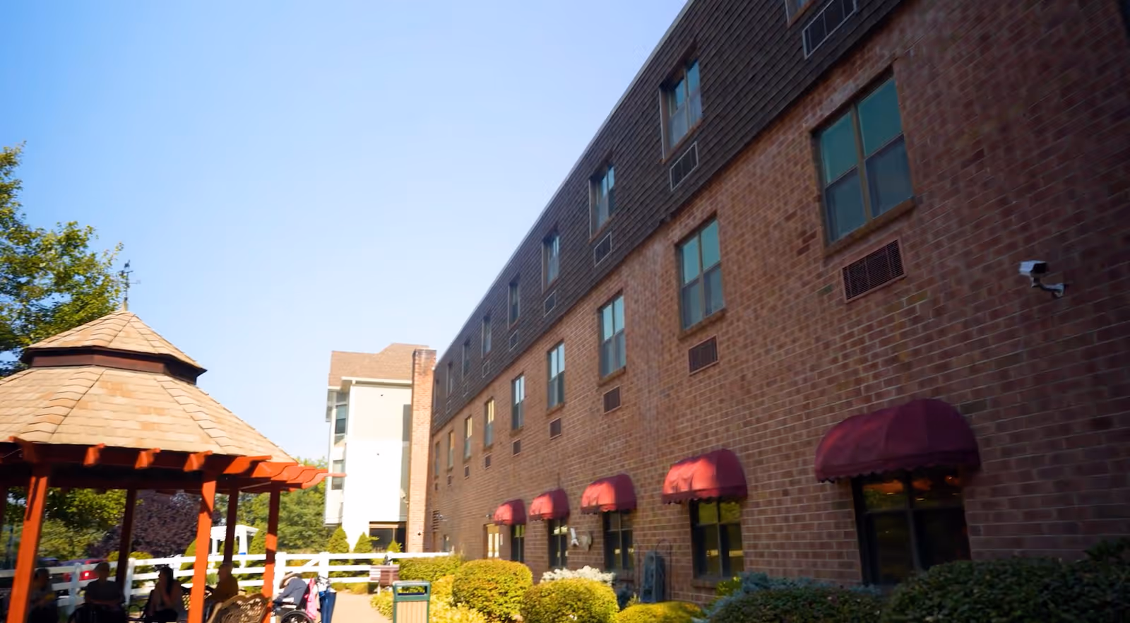 Exterior view of a multi-story brick building with several windows, some with red awnings. In the foreground, there is a wooden gazebo with a shingled roof and some outdoor seating. The scene is set on a clear, sunny day with blue skies and surrounding greenery.
