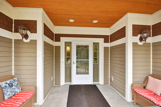 Covered entrance area with a white door featuring glass panels, flanked by two wicker chairs with red cushions and decorative pillows. The walls are beige with white trim and brown accents, and the ceiling is wooden with recessed lighting.
