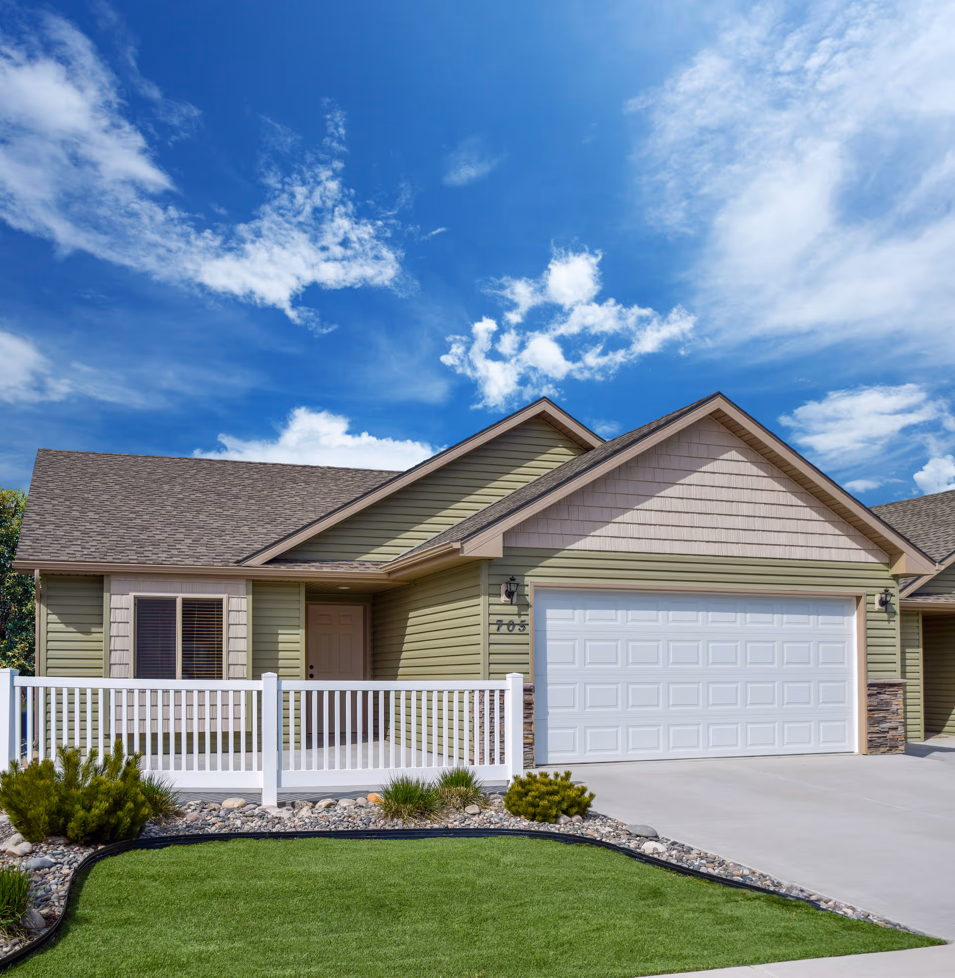 Exterior view of a single-story residential-style building with light green siding, a white garage door, a small front porch with white railing, and a well-maintained lawn and landscaping under a partly cloudy blue sky.