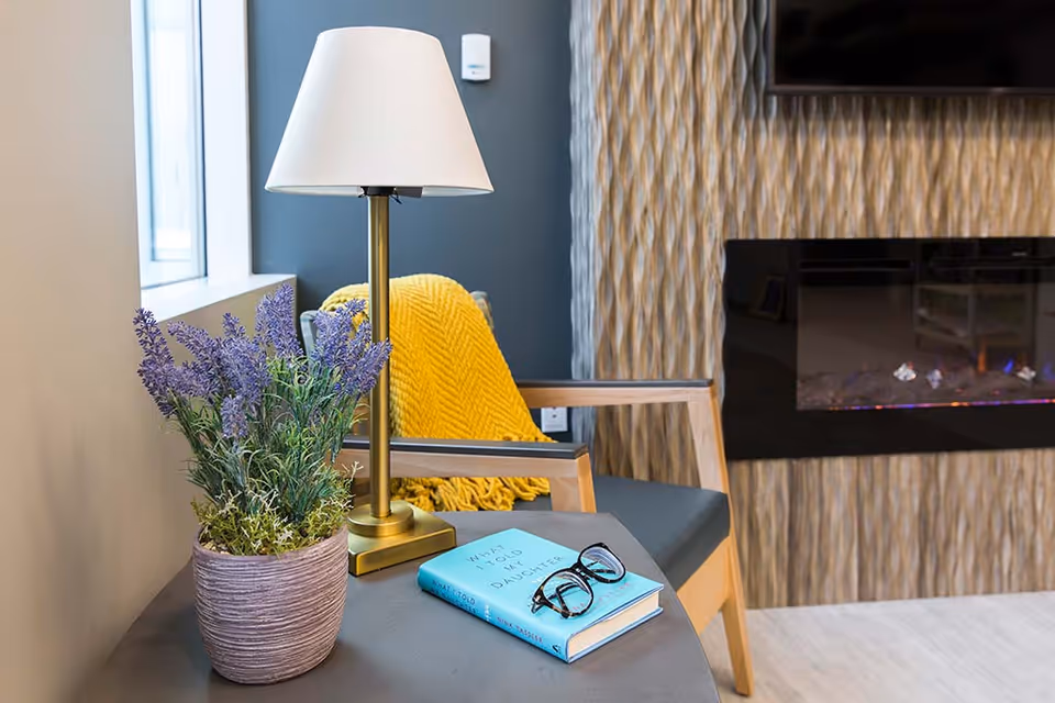 Cozy seating area with a table holding a lamp, potted lavender, a book and glasses beside a chair with a yellow throw in front of a wall-mounted electric fireplace.