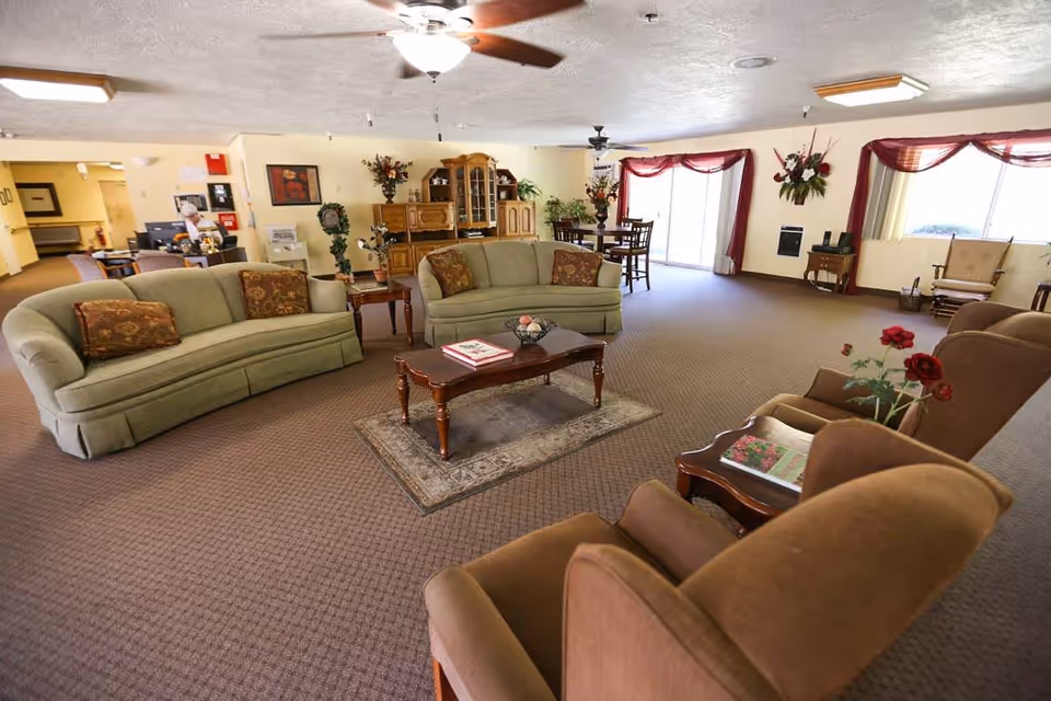 A spacious living room area in an assisted living facility with two green sofas, two brown armchairs, a wooden coffee table on a small rug, and a wooden cabinet with decorative items. There is a dining table with chairs near large windows with red curtains, ceiling fans, and various plants and flowers decorating the room.