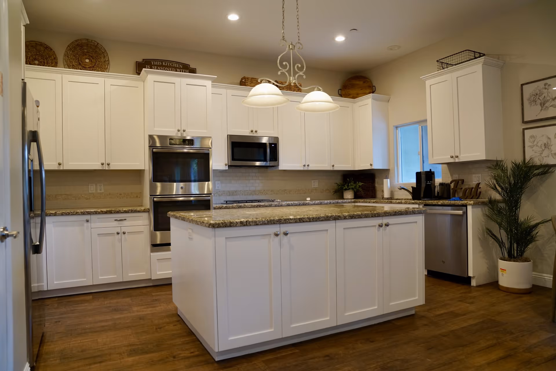 A modern kitchen with white cabinets, granite countertops, and stainless steel appliances including a double oven, microwave, refrigerator, and dishwasher. There is a large kitchen island in the center with matching cabinetry and granite top. The floor is wooden, and the room is well-lit with recessed ceiling lights and two hanging pendant lights above the island. Decorative items such as woven baskets, framed artwork, a potted plant, and a sign that reads 'Eat' are visible.