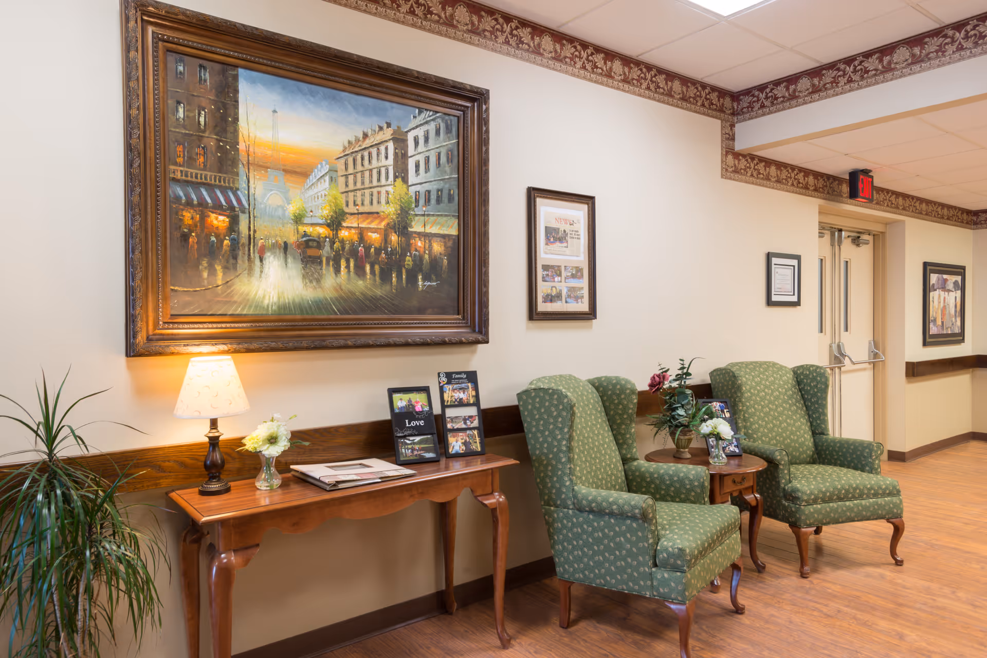 A cozy senior facility sitting area with two green upholstered armchairs, a small side table, a console table with a lamp and framed artwork on the wall.