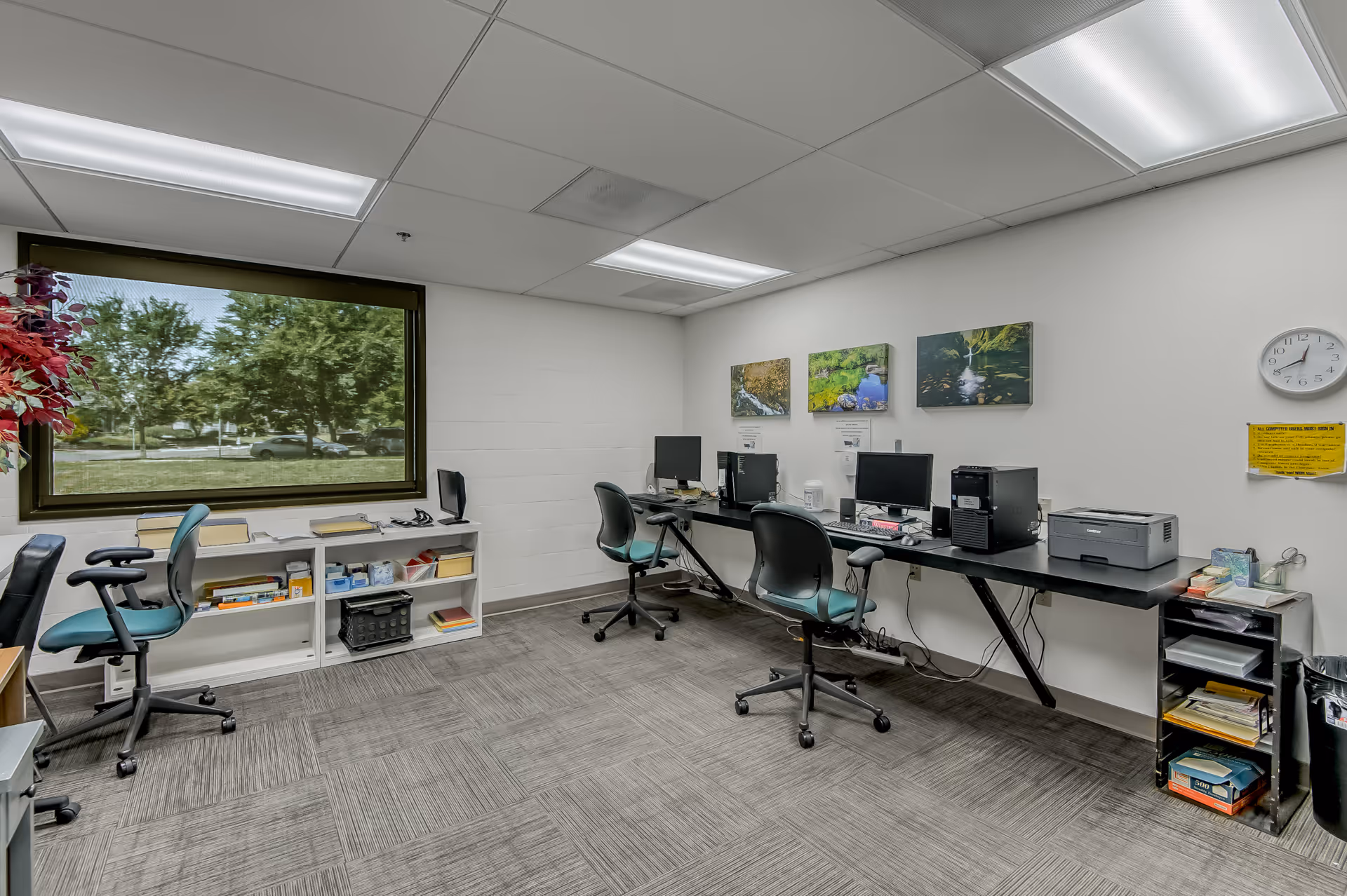 Small computer/workroom with a row of desks, office chairs, computers and a large window showing trees outside.