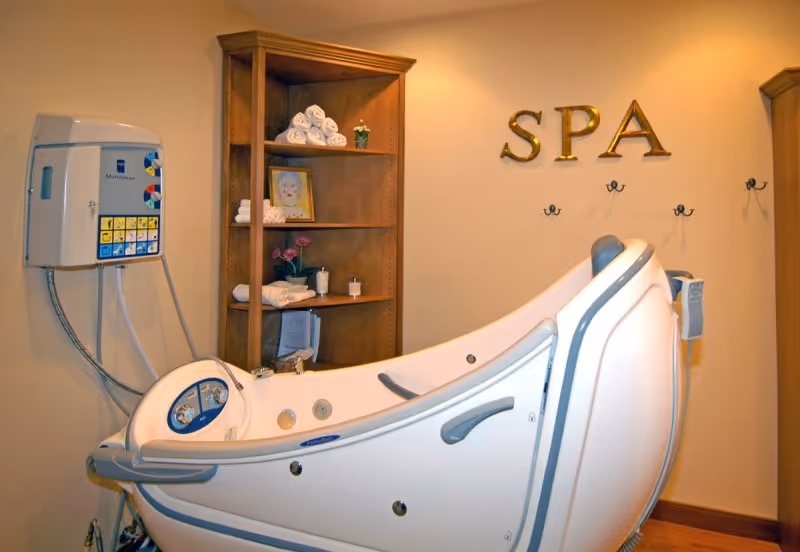 A spa room featuring a white hydrotherapy tub, wooden shelving with towels and decor, and gold letters spelling 'SPA' on the wall.