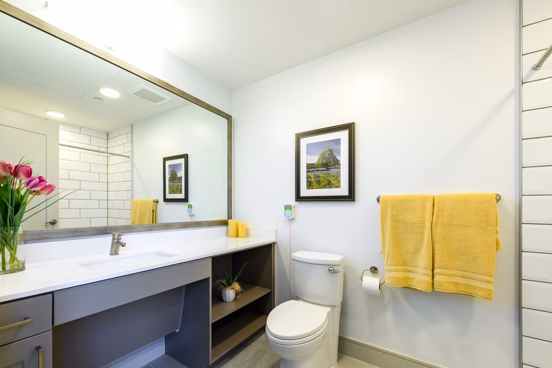 A clean and modern bathroom featuring a large mirror above a white countertop with a built-in sink. There is a vase with pink tulips on the left side of the counter. Below the counter, there is open shelving with a small potted plant. On the right side of the image, a white toilet is positioned against the wall, with a toilet paper holder and two yellow towels hanging on a towel rack above it. A framed picture hangs on the wall above the toilet. The shower area with white subway tiles and a glass door is visible in the mirror reflection.