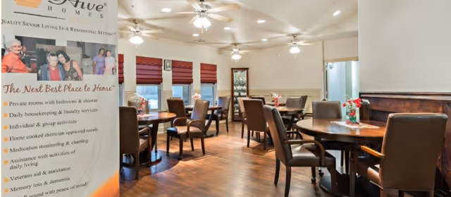 Interior view of a senior living facility dining room with multiple round tables and brown leather chairs. The room has wooden flooring, ceiling fans with lights, and windows with red blinds. A promotional banner for BeeHive Homes is visible on the left side of the image.