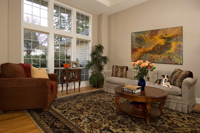 Sunny living room with large floor-to-ceiling windows, two sofas, a coffee table on an ornate rug, potted plants, wall art and a small dog on the couch.