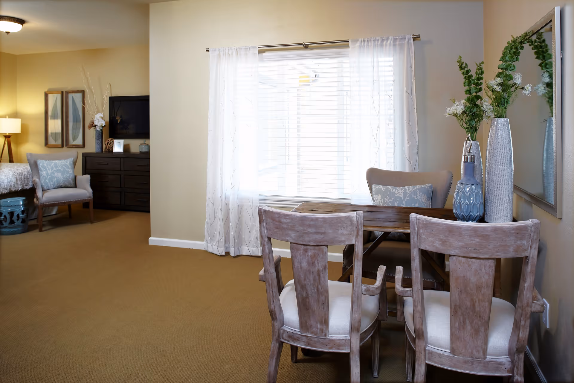 A cozy interior room with a small wooden dining table and two chairs in the foreground. A window with sheer white curtains lets in natural light. To the left, there is a sitting area with an upholstered chair, a dark wooden dresser with a TV on top, and part of a bed with a lamp beside it. Decorative vases with green plants and flowers are placed on the table and dresser.