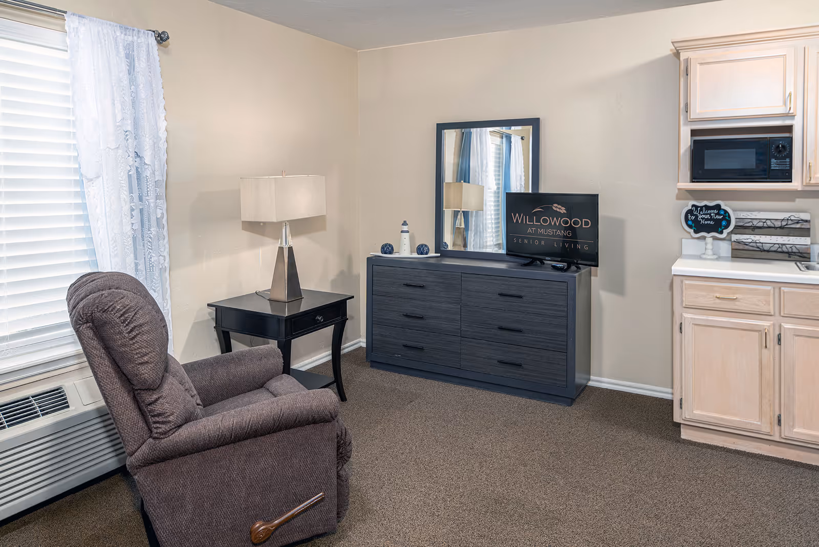 A senior living studio with a recliner, side table and lamp, a dresser with mirror and TV, and a small kitchenette area.