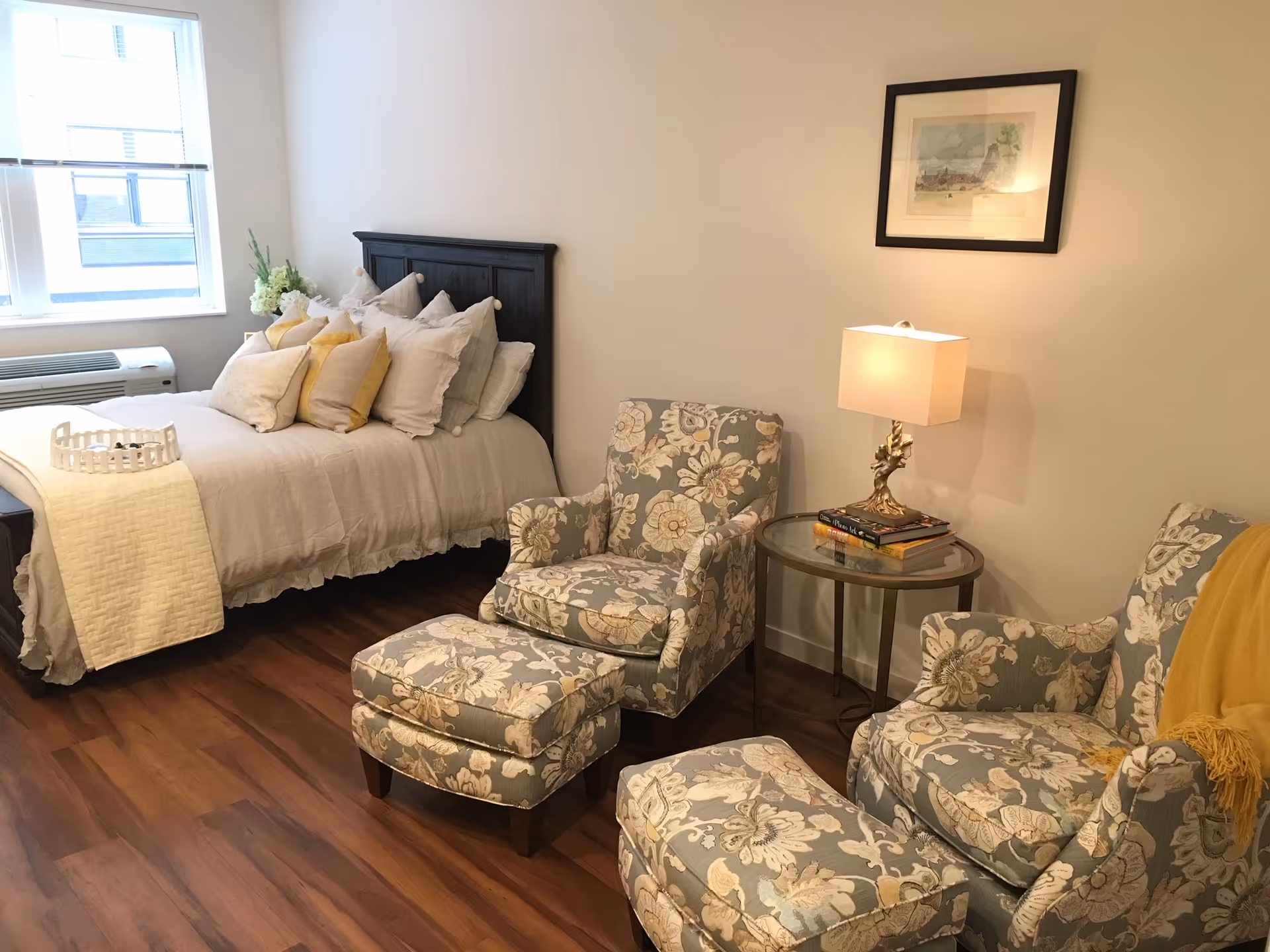 A cozy bedroom with a bed by the window and two floral armchairs with ottomans arranged around a side table and lamp.