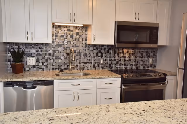 Modern kitchen with white cabinets, a mosaic tile backsplash in shades of gray and black, a stainless steel dishwasher, sink with a curved faucet, stainless steel oven with a black microwave above it, and a granite countertop.
