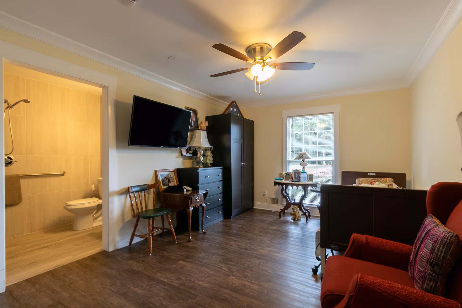 A cozy senior living room with wooden flooring, a ceiling fan with lights, a mounted flat-screen TV, a black dresser, a wooden chair, a small table with a lamp and framed photos, a window with blinds, a red armchair with a decorative pillow, and a view into an adjacent bathroom with a toilet and grab bars.