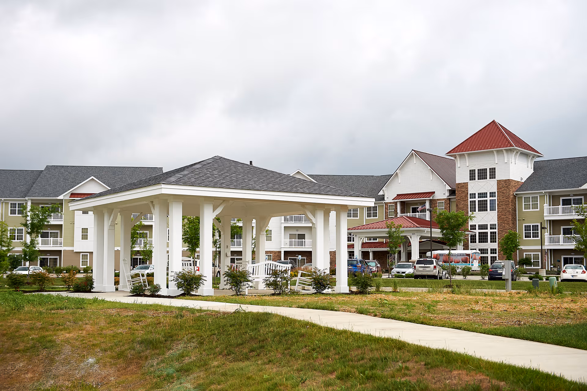 White gazebo and walkway in front of a multi-story senior living building with red roofs and parked cars.