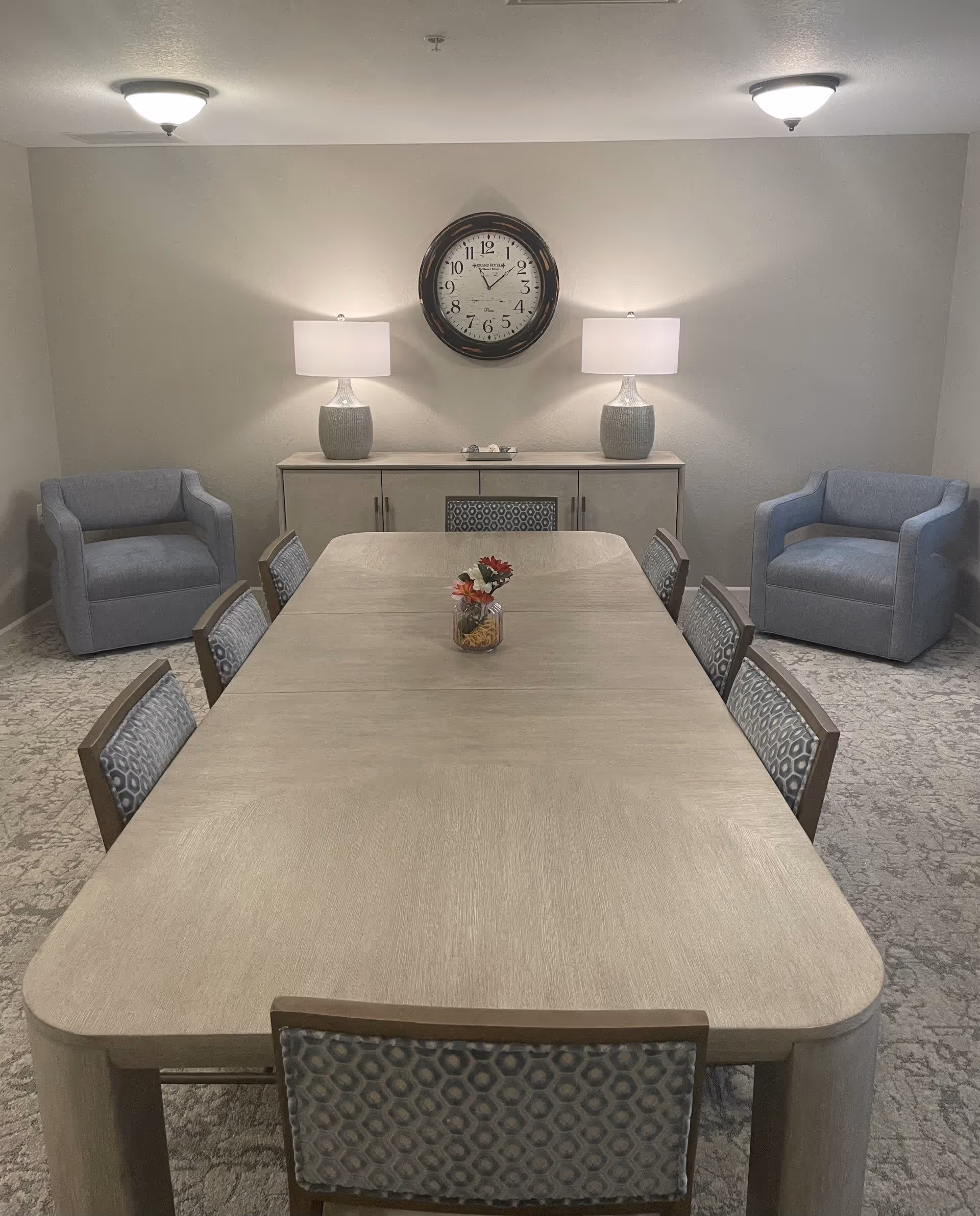 A dining room with a long rectangular wooden table surrounded by eight chairs with patterned upholstery. At the far end of the room, there is a sideboard with two table lamps and a large round wall clock above it. Two blue armchairs are positioned against the walls on either side of the sideboard. The room has neutral-colored walls and carpeted flooring.