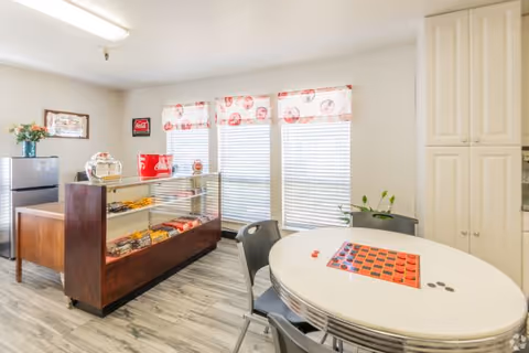 A bright room with large windows covered by white blinds and red patterned valances. There is a round white table with a checkerboard game set up and four black chairs around it. To the left, there is a glass display case containing snacks and a small refrigerator with flowers on top. The floor has a light wood pattern, and white cabinets are visible on the right side of the image.