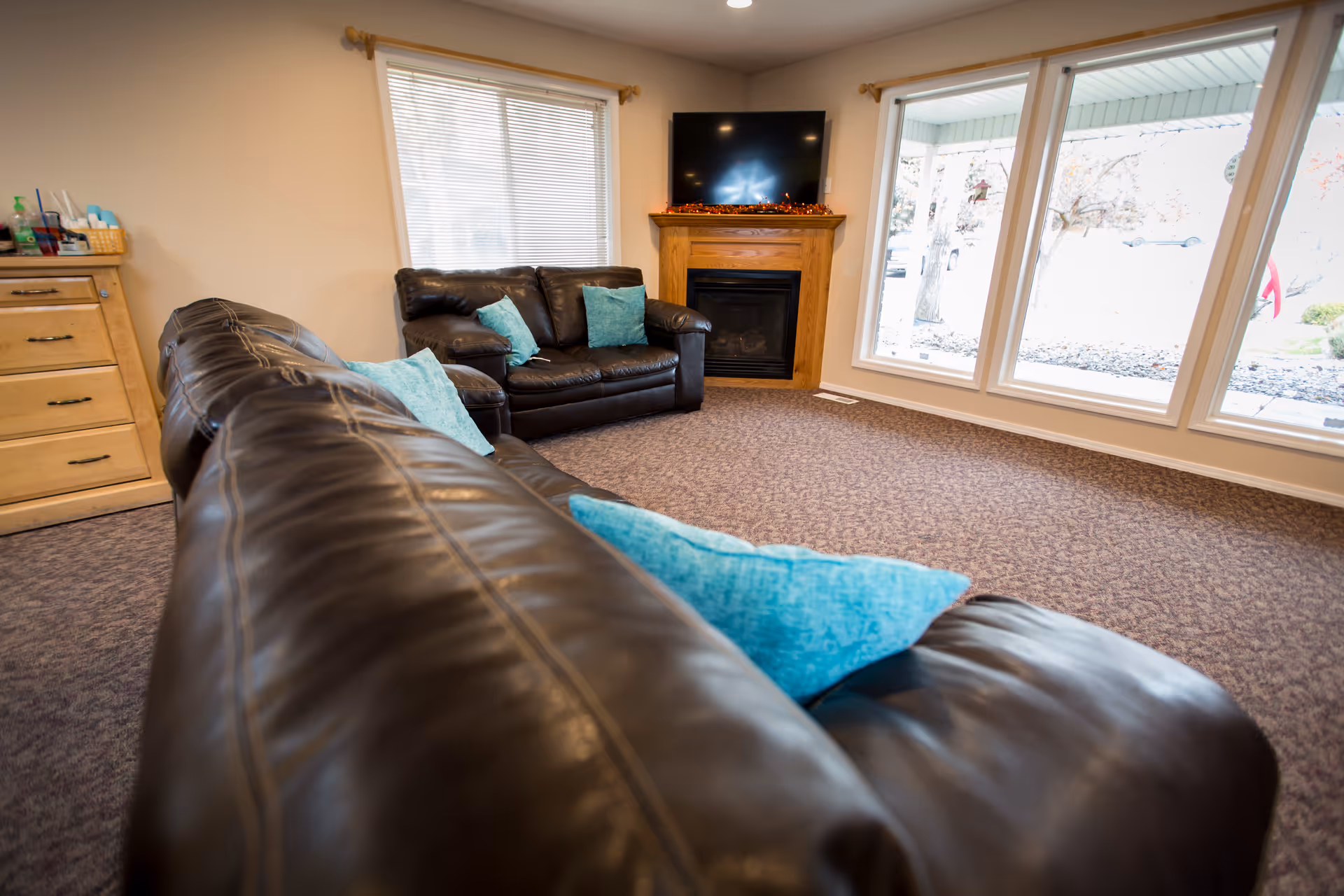 Cozy assisted-living lounge with brown leather sofas and turquoise pillows, a fireplace with TV above, large windows, and carpeted floor.