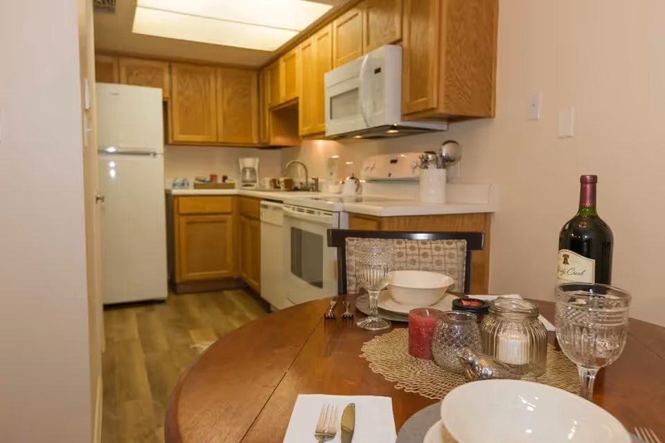 A cozy kitchen and dining area with wooden cabinets, a white refrigerator, stove, microwave, and dishwasher. The dining table is set with plates, bowls, glasses, cutlery, candles, and a bottle of wine.