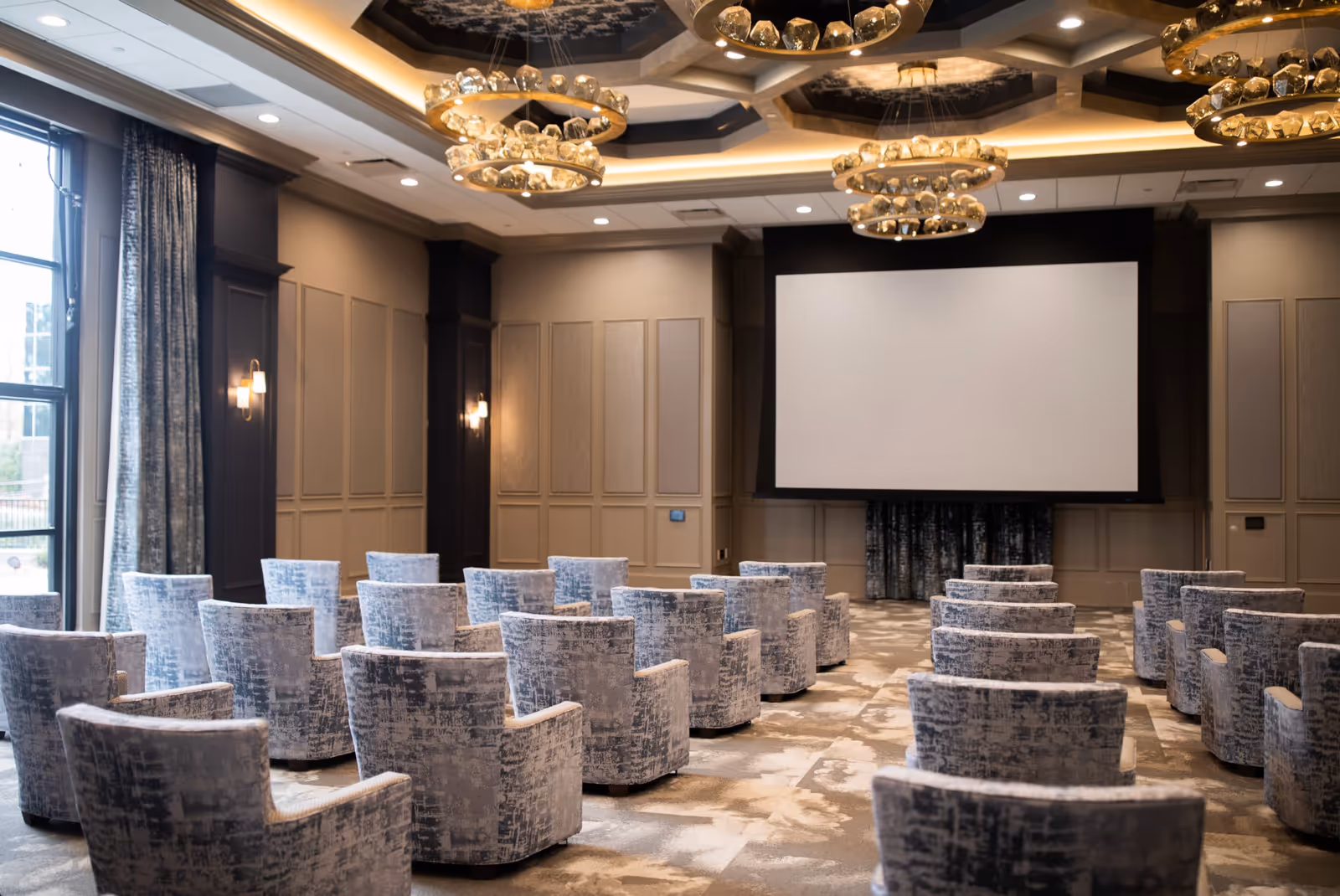 A modern conference or presentation room with rows of upholstered armchairs facing a large blank projection screen. The room features elegant chandeliers, paneled walls, and large windows with curtains allowing natural light to enter.