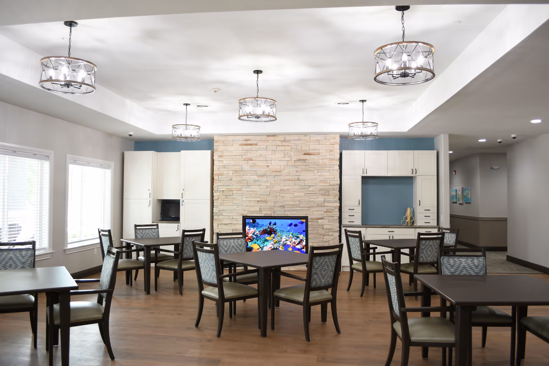 Well-lit communal dining area with tables and chairs, pendant lights, a stone accent wall with a television displaying an aquarium, and cabinetry.
