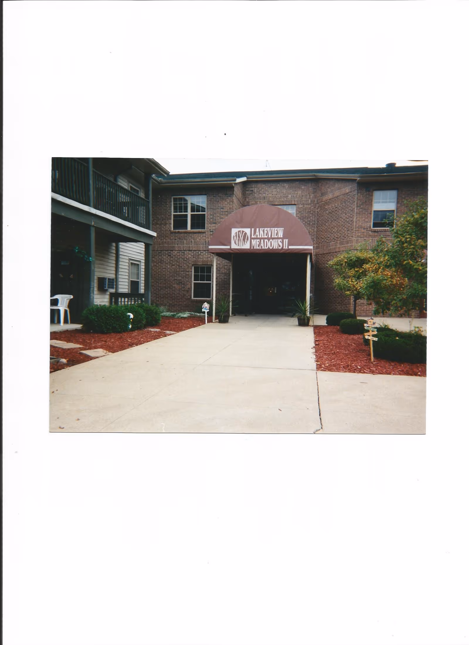 Entrance to a brick building with a maroon awning that reads 'Lakeview Meadows II'. The entrance is flanked by two potted plants and there are landscaped areas with bushes and mulch on either side of the concrete walkway leading to the door.