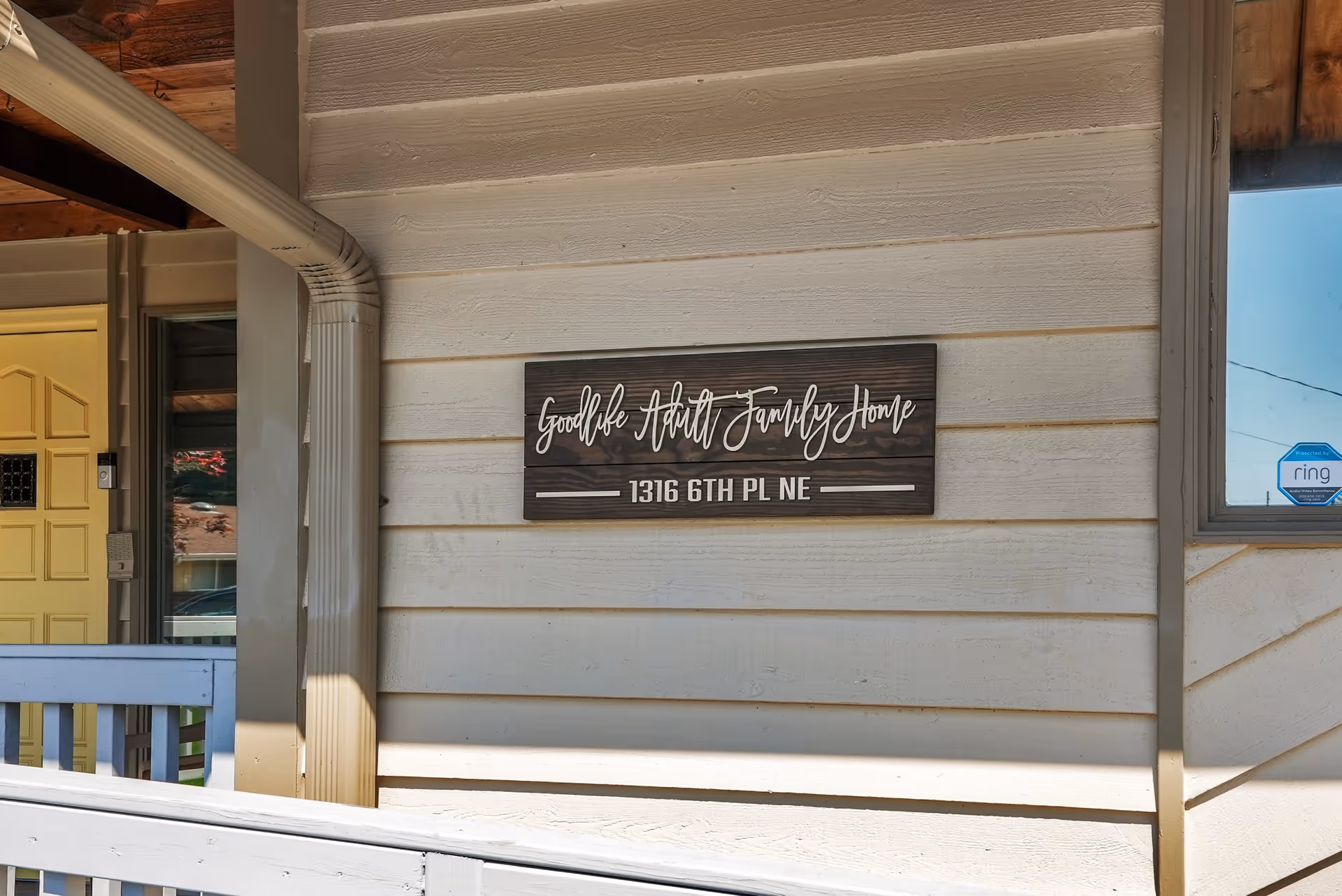 Exterior wall of a building with a wooden sign that reads 'Goodlife Adult Family Home 1316 6TH PL NE'. The wall is light-colored horizontal siding, and there is a yellow door and a window with a security sticker visible.