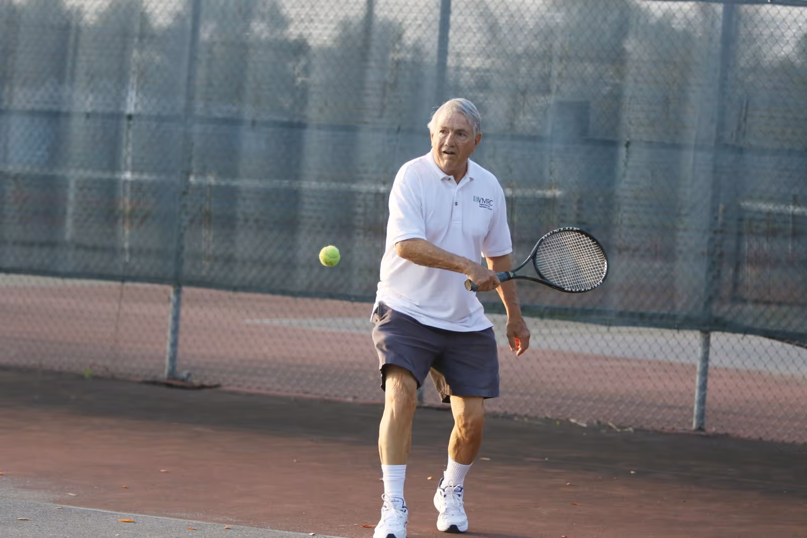 An elderly man playing tennis on an outdoor tennis court, wearing a white polo shirt, dark shorts, white socks, and white sneakers, holding a tennis racket and preparing to hit a tennis ball.