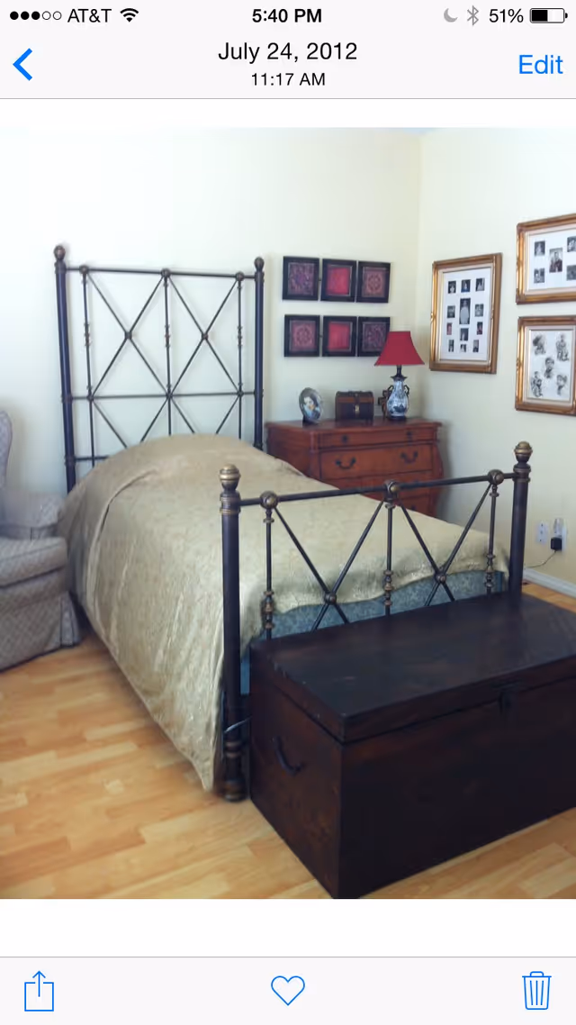 A bedroom with a single metal-framed bed covered with a beige bedspread. Next to the bed is a wooden chest at the foot and a wooden dresser with a red lamp, decorative items, and framed pictures on the wall above it. There is also a cushioned armchair partially visible on the left side. The floor is light-colored wood.