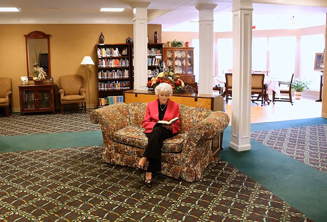 An elderly woman with white curly hair wearing a red jacket and black pants is sitting on a floral-patterned couch reading a book in a spacious room. The room has patterned carpet, white columns, bookshelves filled with books, armchairs, a wooden cabinet with glass doors, and a dining area with tables and chairs in the background near large windows letting in natural light.