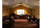 Interior view of a spacious common area with multiple tables and chairs arranged on a patterned carpet. The room has a high vaulted ceiling with recessed lighting and a large window at the far end letting in natural light. There is a piano near the window and the walls are painted a warm beige color.