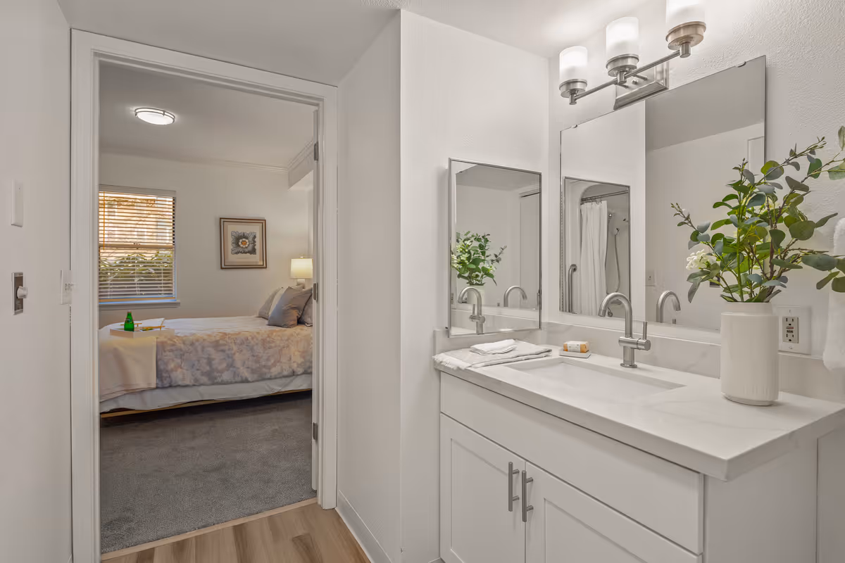 A bright and clean bathroom with a white vanity, dual sinks, and a large mirror. A vase with green foliage is placed on the countertop. Through an open door, a bedroom with a bed, pillows, a framed picture on the wall, and a window with blinds is visible.