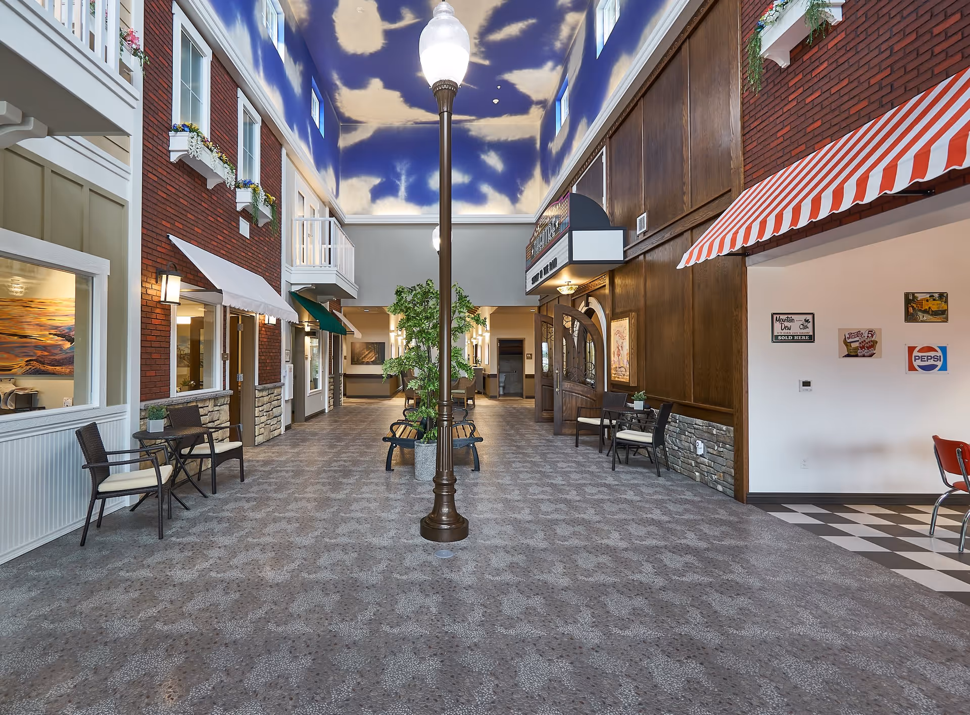 Indoor hallway designed to resemble an outdoor street scene with a patterned floor, a streetlamp in the center, benches, potted plants, and storefront-style windows and awnings on either side. The ceiling is painted blue with white clouds, and there are decorative signs and posters on the walls.