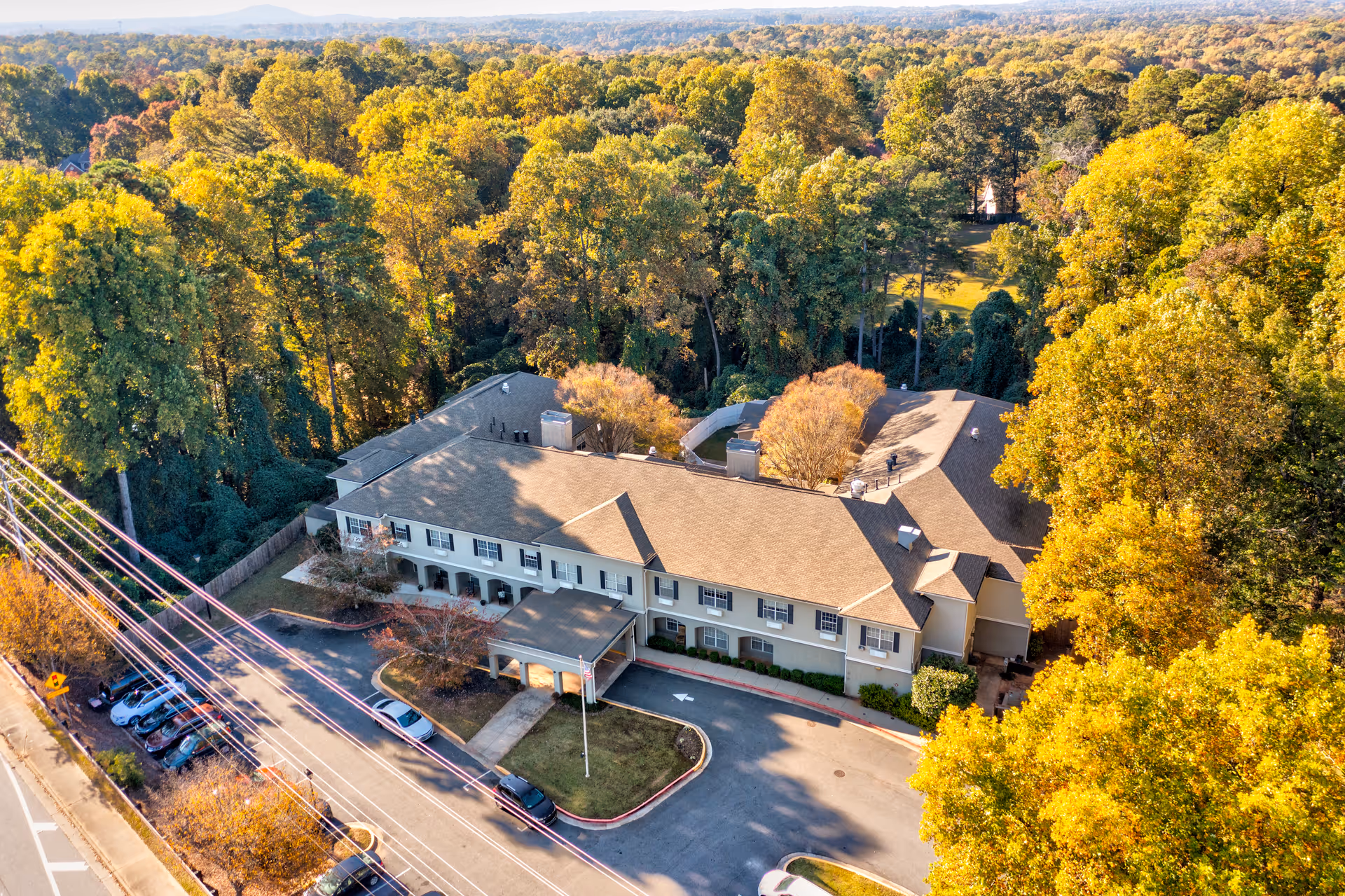 Aerial view of Greenwood Place Assisted Living and Memory Care building surrounded by dense trees with autumn foliage. The building has a beige exterior with a brown roof and a covered entrance. Several cars are parked along the driveway and street adjacent to the facility.