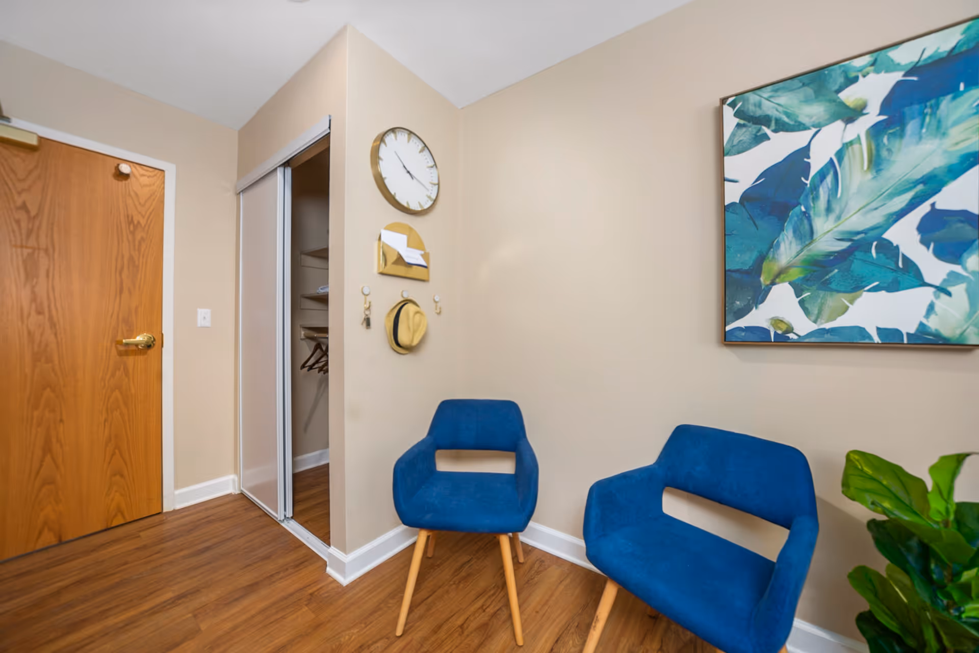 Interior corner of a room with two blue chairs, a wooden door, a closet with sliding doors, a wall clock, a small wall-mounted mail holder, hooks with a hat and keys, a large framed artwork with green and blue leaves, and a green plant in the corner.