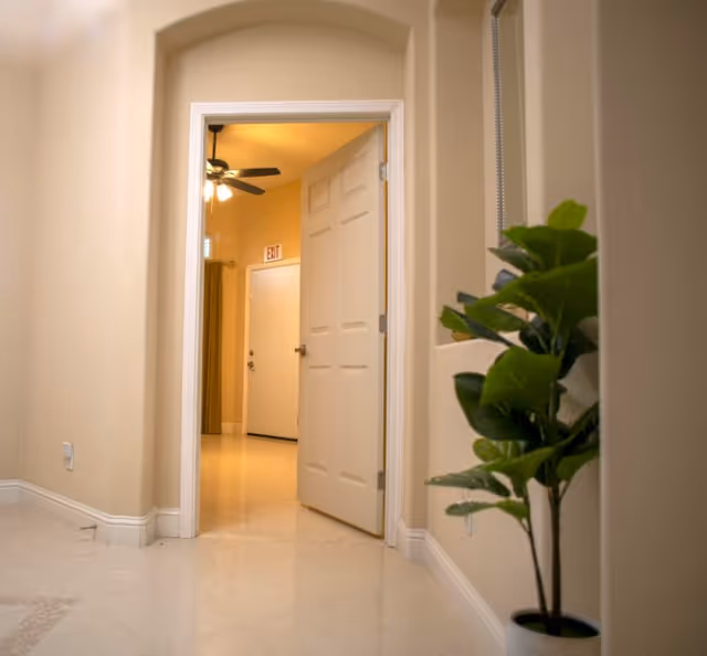View of an open door in a hallway with beige walls and tiled floor, a potted plant on the right side, and a ceiling fan with lights visible in the adjacent room.